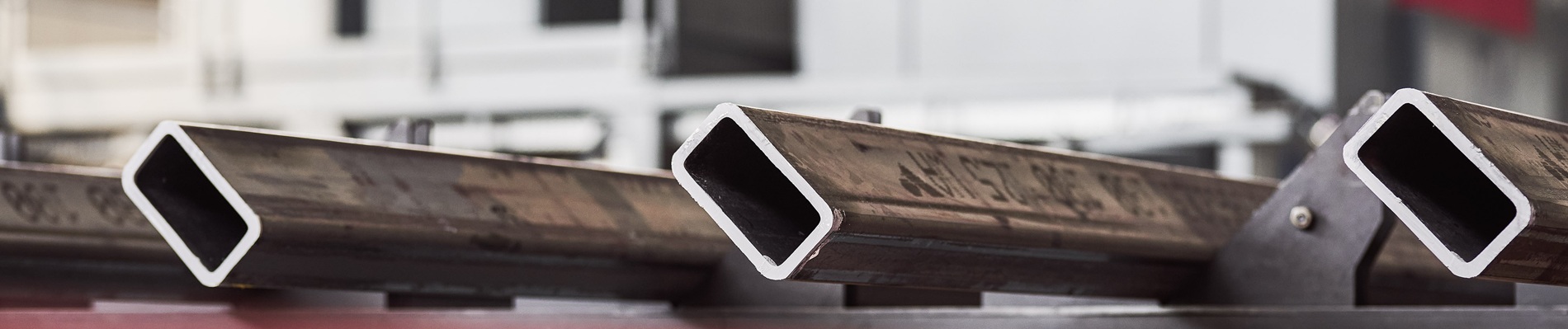 Nahaufnahme mehrerer rechteckiger Metallrohre mit hohlen Innenräumen, die parallel zueinander ausgerichtet sind, vor einem unscharfen industriellen Hintergrund.