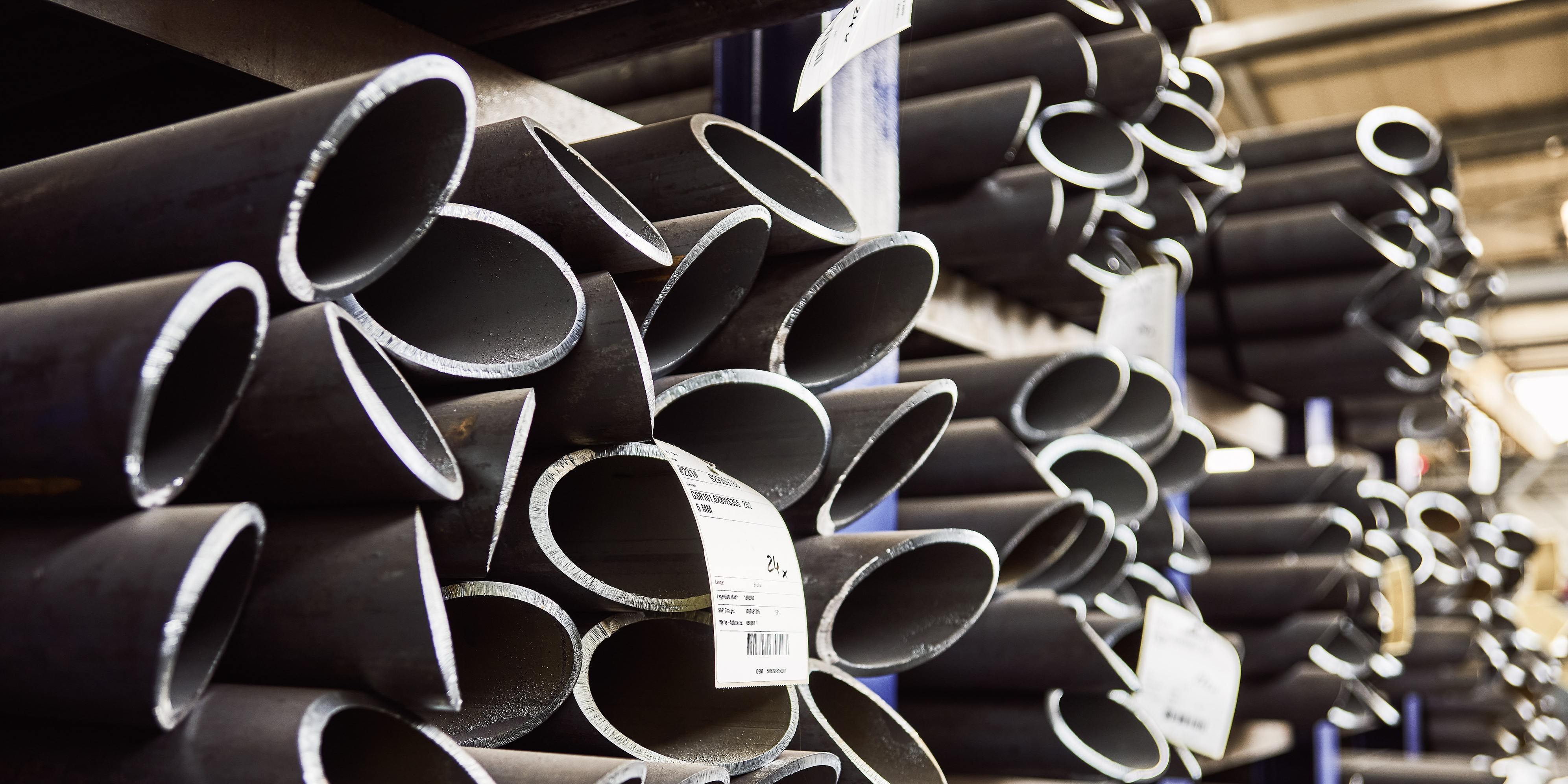 Stapel von industriellen Metallrohren mit Etiketten, angeordnet in einer Lagerhalle, die ihre hohlen zylindrischen Formen zur Geltung bringen.
